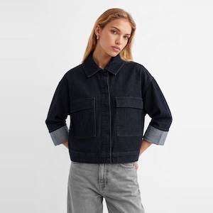 May include: A dark indigo denim jacket featuring a classic collar, a button-front closure, and two large rectangular chest pockets. The jacket has long sleeves with wide, folded cuffs that reveal a lighter blue interior lining and a cropped hem.