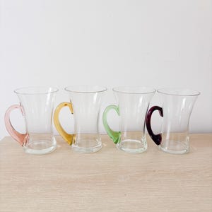 May include: Four clear glass mugs with a flared rim and a rounded base. Each mug features a distinctively coloured handle in pink, yellow, light green, and dark purple. The mugs are arranged in a horizontal row, showcasing their transparent bodies.