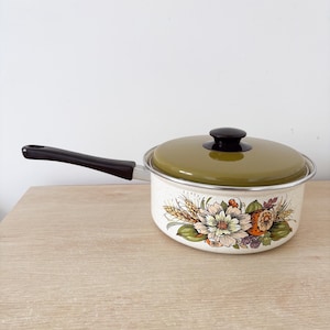 May include: A vintage enamel saucepan with a lid. The pan is off-white with a floral design in orange, green, and cream. It has a black handle and a green lid with a black knob. A classic piece of kitchenware.