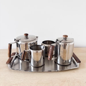 Op de afbeelding: Een roestvrijstalen theeservies bestaande uit een theepot, een koffiepot, een melkkan en een open suikerpot, allemaal voorzien van donkerhouten handgrepen en dekselknoppen, geplaatst op een bijpassend rechthoekig roestvrijstalen dienblad.