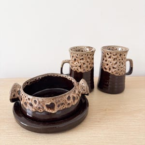 May include: A set of vintage ceramic tableware with a dark brown glaze and speckled tan rims. The set includes two mugs and a bowl with handles, resting on a matching saucer. The pieces are arranged on a light wood surface.