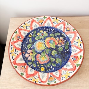 Op de afbeelding: Een ronde, handbeschilderde keramische plaat met een bloemenontwerp. De plaat heeft een diepblauwe kern met kleurrijke bloemen en groene bladeren. De rand heeft een rode rand en een herhalend patroon van rode en witte vormen.