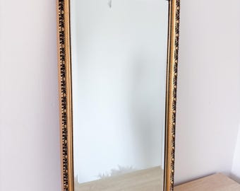 May include: A rectangular wall mirror featuring a decorative gold-toned frame with a repeating circular and linear pattern. The mirror has a clear, reflective glass surface and is positioned at an angle, showcasing the detailed molding of the frame.