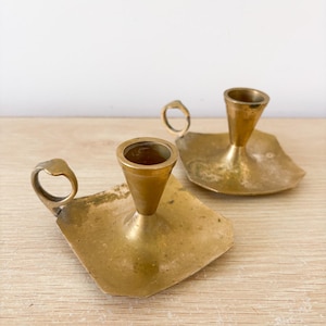 Peut inclure: Deux bougeoirs vintage en laiton. Chacun a une base carrée avec une poignée et une coupelle conique pour tenir une bougie. Les bougeoirs ont un aspect terni et vieilli, suggérant qu'ils sont antiques ou vintage.