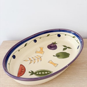 Può includere: Piatto da portata ovale in ceramica con interno color crema e bordo viola e blu. Il piatto è decorato con illustrazioni colorate di verdure e pasta, tra cui pomodori, melanzane e piselli.