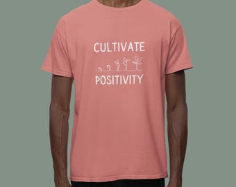 Pode incluir: Uma camiseta rosa coral com o texto "Cultivate Positivity" em branco. O texto é acompanhado por um desenho de linha de cinco plantas crescendo.