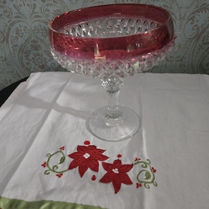 May include: A clear glass candy dish with a red rim and diamond-shaped pattern. The dish is on a white tablecloth with red and green embroidery.