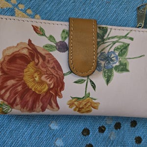 May include: A light pink leather wallet with a floral design. The wallet features a large red and yellow flower, along with other flowers and green leaves. It has a brown leather tab closure and a silver zipper.