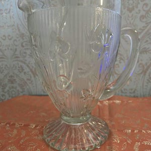 May include: A clear glass pitcher with a textured surface and a floral design. The pitcher has a wide base and a handle.