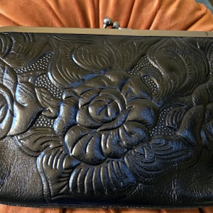 May include: Black leather clutch with a floral embossed design. The clutch features a kiss-lock closure and a decorative metal frame. The embossed pattern includes roses and leaves, adding a touch of elegance to the accessory.