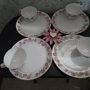 May include: Four white teacups and saucers with pink floral designs and gold trim are arranged on a black surface. A pink metal flower decoration with a heart in the center is also present. The teacups and saucers are a vintage tea set.