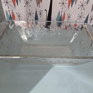 May include: A clear, rectangular glass baking dish with a textured floral pattern. The dish has a slightly raised rim and is designed for oven use. The glass appears to be thick and durable, suitable for baking various dishes.