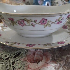 May include: A white porcelain gravy boat with a gold trim and a floral design featuring pink flowers and green leaves. The boat is sitting on a matching saucer.