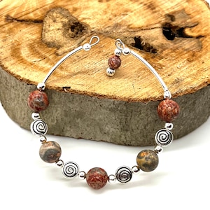 Può includere: Un braccialetto rigido in argento con un ciondolo a spirale in argento e perline di diaspro rosso, marrone e grigio.