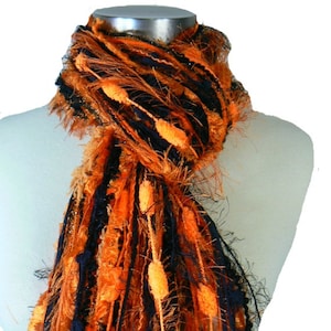 May include: A long, black and orange scarf with fuzzy texture and orange pom-poms. The scarf is draped over a white mannequin.