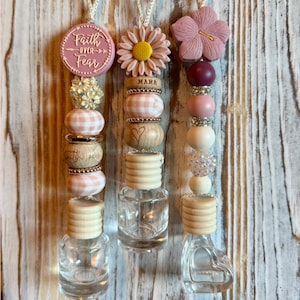 May include: Three decorative car air fresheners with hanging strings. Each features a glass bottle, wooden beads, and decorative accents like flowers and the text "Faith Over Fear" and "Mama". The background is a wooden surface.