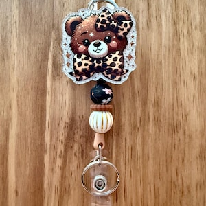 Puede incluir: Un portatarjetas con un oso de dibujos animados con un lazo y detalles de estampado de leopardo. El portatarjetas tiene un diseño de cuentas con una cuenta floral negra, una cuenta a rayas blancas y doradas y una cuenta rosa claro. Tiene un clip de plástico transparente y una cinta blanca.