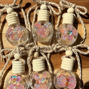 Puede incluir: Seis ambientadores para coche de cristal transparente con tapas de madera y formas decorativas rellenas de purpurina de colores. Cada botella está suspendida por una cuerda beige. Las formas incluyen un lazo, una mariposa y una flor.