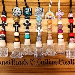 Könnte beinhalten: Eine Sammlung von Auto-Lufterfrischern mit dekorativen Perlen und religiösen Anhängern. Jeder Lufterfrischer hat eine kleine Glasflasche und einen Holzdeckel. Die Anhänger zeigen Kreuze, die Worte "Pray", "Love Jesus" und "God" sowie das Wort "Blessed."