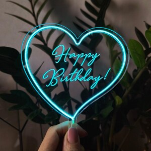 Könnte beinhalten: Ein transparenter, herzförmiger Tortendekor aus Acryl mit einem neongrünen Umriss und dem Text "Happy Birthday!" im Inneren.