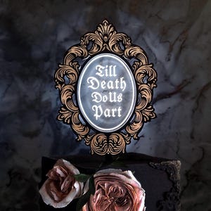 Op de afbeelding: Een zwarte en gouden taarttopper met een sierlijk frame en de tekst "Till Death Do Us Part" in witte letters.