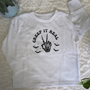 May include: White long-sleeved shirt with a black skeleton hand making a peace sign and the text "CREEP IT REAL" in a circle around the hand. There are also black bats and stars around the hand.