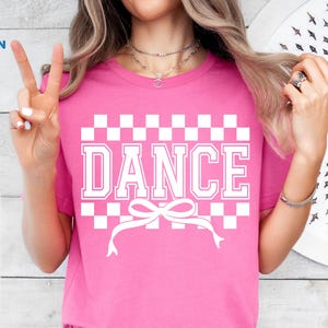 Pode incluir: Camiseta rosa com um padrão xadrez branco e a palavra "DANCE" em letras brancas. Um laço branco está centralizado abaixo da palavra.