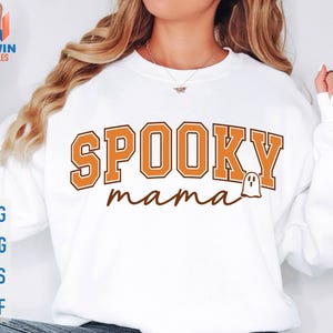 May include: White sweatshirt with the text "SPOOKY mama" in orange and brown lettering, featuring a small ghost graphic. The sweatshirt also has the words "SVG, PNG, EPS, DXF" in blue.