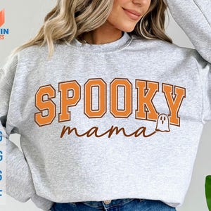 May include: A light gray sweatshirt with the words "SPOOKY mama" in orange and brown lettering. A small ghost graphic is next to the word "mama". The sweatshirt also has the words "SVG, PNG, EPS, DXF" in a vertical column.
