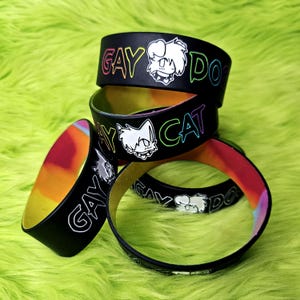 May include: Black rubber bracelets with the words "GAY DOG" and "GAY CAT" in rainbow lettering. Each bracelet features a white cartoon animal illustration. The inner band has a colorful iridescent finish.