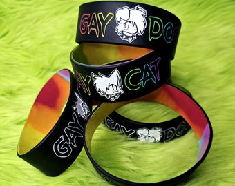 May include: Black rubber bracelets with the words "GAY DOG" and "GAY CAT" in rainbow lettering. Each bracelet features a white cartoon animal illustration. The inner band has a colorful iridescent finish.