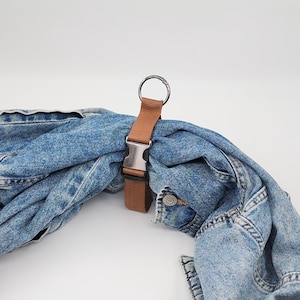 Könnte beinhalten: Ein brauner Riemen mit einer silbernen Schnalle und einem Metallring ist an einer blauen Jeansjacke befestigt. Die Jacke ist gefaltet und der Riemen wird verwendet, um sie zusammenzuhalten. Der Hintergrund ist schlicht weiß.