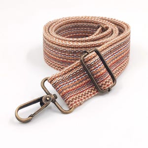 May include: A woven bag strap featuring a multi-coloured striped pattern in shades of orange, brown, white, and blue. The strap includes an adjustable antique brass-toned metal slider and a matching swivel lobster clasp hook at the end.
