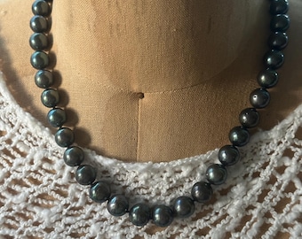 Op de afbeelding: Een ketting van donkergrijze parels, tentoongesteld op een mannequin. De parels zijn rond en gelijkmatig van formaat, met een subtiele glans. De ketting is gedrapeerd over een wit, open geweven kledingstuk.