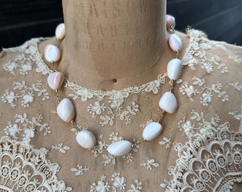 May include: A necklace featuring large, oval-shaped, white and pink stones strung together with gold-colored links. The necklace is displayed on a mannequin with a tan fabric and a vintage lace overlay.