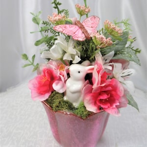May include: A decorative arrangement in a pink glass vase featuring a small white ceramic rabbit figurine nestled in green moss, surrounded by artificial pink and white flowers, green foliage, and a pink butterfly ornament perched on top.