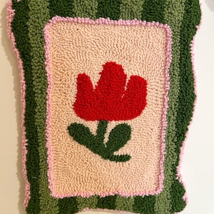 Pode incluir: Um tapete retangular feito &agrave; m&atilde;o com um desenho de tulipa vermelha. O tapete apresenta um centro bege claro, uma borda rosa e uma borda externa verde listrada. A tulipa tem folhas verdes. O tapete tem aproximadamente 45 cm de largura e 61 cm de comprimento.