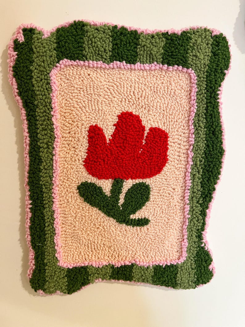 Pode incluir: Um tapete retangular feito &agrave; m&atilde;o com um desenho de tulipa vermelha. O tapete apresenta um centro bege claro, uma borda rosa e uma borda externa verde listrada. A tulipa tem folhas verdes. O tapete tem aproximadamente 45 cm de largura e 61 cm de comprimento.