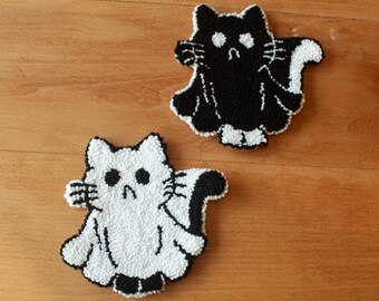 May include: Two cat-shaped coasters, one black and one white, sit on a wooden surface. The coasters have a fluffy, textured appearance with black and white details. The cat designs have large eyes and tails.