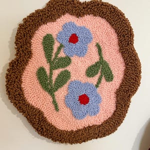 Pode incluir: Uma tape&ccedil;aria de parede redonda e feita &agrave; m&atilde;o com um design floral. A pe&ccedil;a tem um centro rosa claro com duas flores azuis com centros vermelhos e folhas verdes. A borda &eacute; de cor marrom escuro.