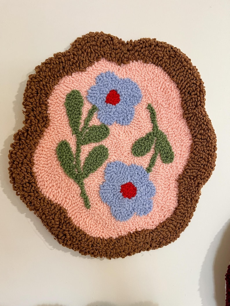Pode incluir: Uma tape&ccedil;aria de parede redonda e feita &agrave; m&atilde;o com um design floral. A pe&ccedil;a tem um centro rosa claro com duas flores azuis com centros vermelhos e folhas verdes. A borda &eacute; de cor marrom escuro.