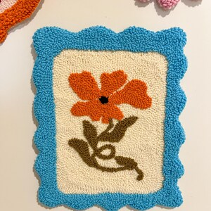 Pode incluir: Uma tape&ccedil;aria de parede quadrada e texturizada com uma borda azul ondulada. O centro apresenta uma flor laranja com caules e folhas marrons sobre um fundo creme. Outros itens semelhantes s&atilde;o vis&iacute;veis no fundo.