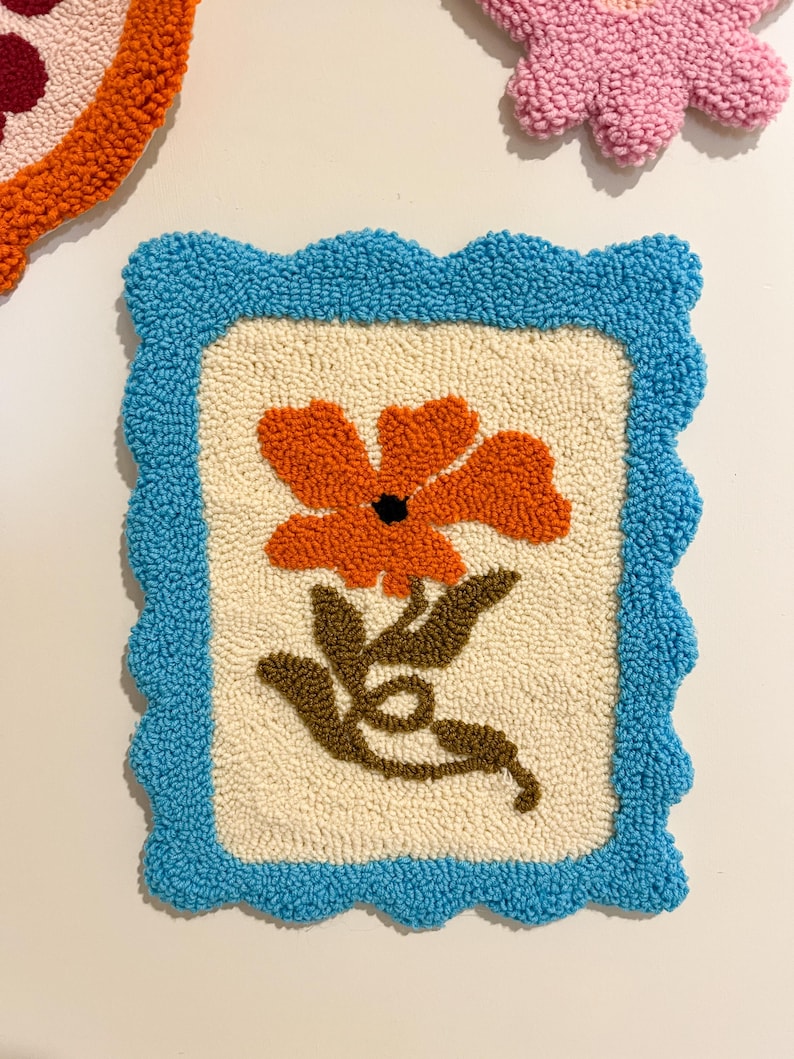 Pode incluir: Uma tape&ccedil;aria de parede quadrada e texturizada com uma borda azul ondulada. O centro apresenta uma flor laranja com caules e folhas marrons sobre um fundo creme. Outros itens semelhantes s&atilde;o vis&iacute;veis no fundo.