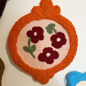 Pode incluir: Uma tape&ccedil;aria decorativa e texturizada com uma borda laranja e um design central com tr&ecirc;s flores bord&ocirc; e folhas verdes sobre um fundo rosa. A tape&ccedil;aria tem uma forma &uacute;nica e ornamentada.