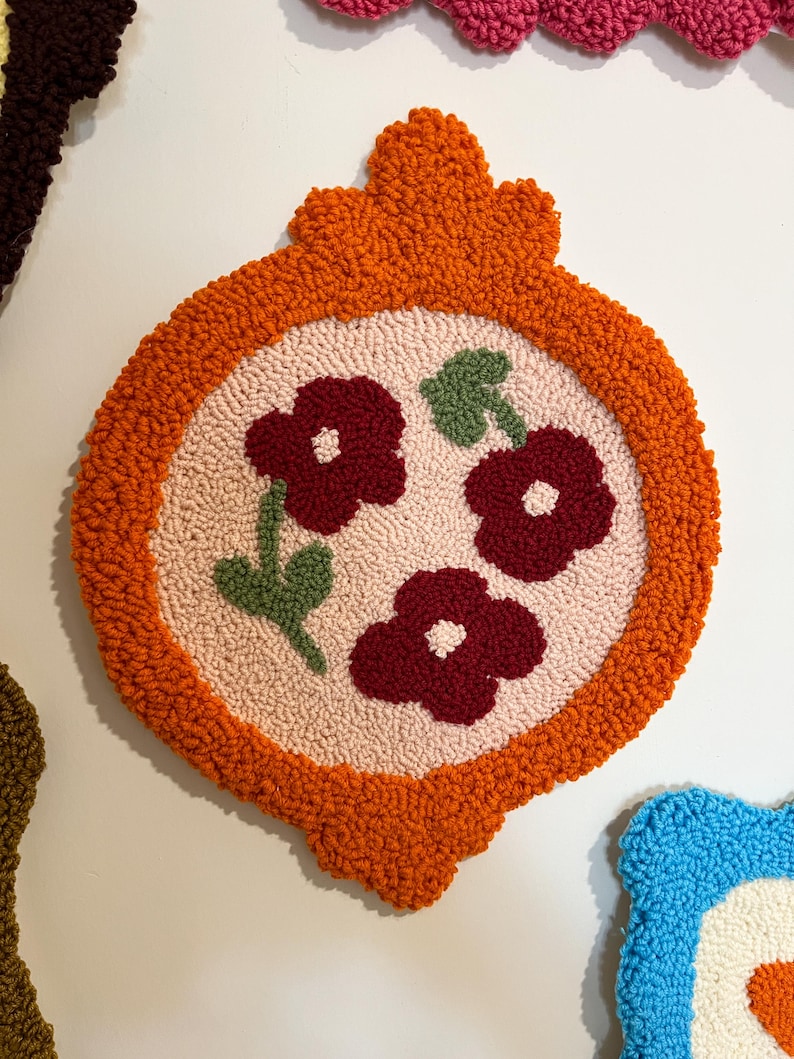 Pode incluir: Uma tape&ccedil;aria decorativa e texturizada com uma borda laranja e um design central com tr&ecirc;s flores bord&ocirc; e folhas verdes sobre um fundo rosa. A tape&ccedil;aria tem uma forma &uacute;nica e ornamentada.
