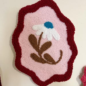 Pode incluir: Uma tape&ccedil;aria de parede texturizada feita &agrave; m&atilde;o com um design de margarida. A obra de arte tem um fundo rosa claro com uma margarida branca, um centro azul e folhas marrons. A borda &eacute; vermelha escura, criando um contraste marcante.
