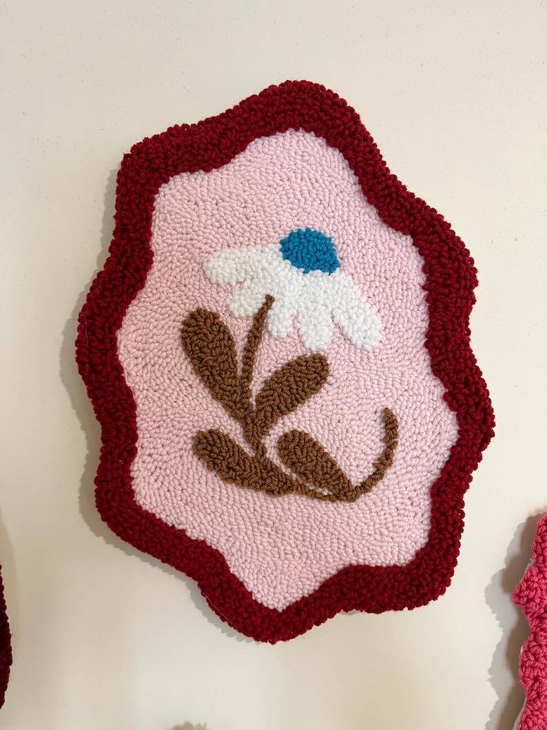 Pode incluir: Uma tape&ccedil;aria de parede texturizada feita &agrave; m&atilde;o com um design de margarida. A obra de arte tem um fundo rosa claro com uma margarida branca, um centro azul e folhas marrons. A borda &eacute; vermelha escura, criando um contraste marcante.