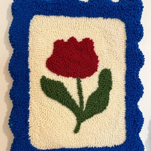 Pode incluir: Uma tape&ccedil;aria de parede texturizada feita &agrave; m&atilde;o com uma tulipa vermelha e folhas verdes sobre um fundo creme. O design &eacute; emoldurado por uma borda ondulada azul brilhante. A obra de arte &eacute; feita com uma t&eacute;cnica de fio em la&ccedil;o.