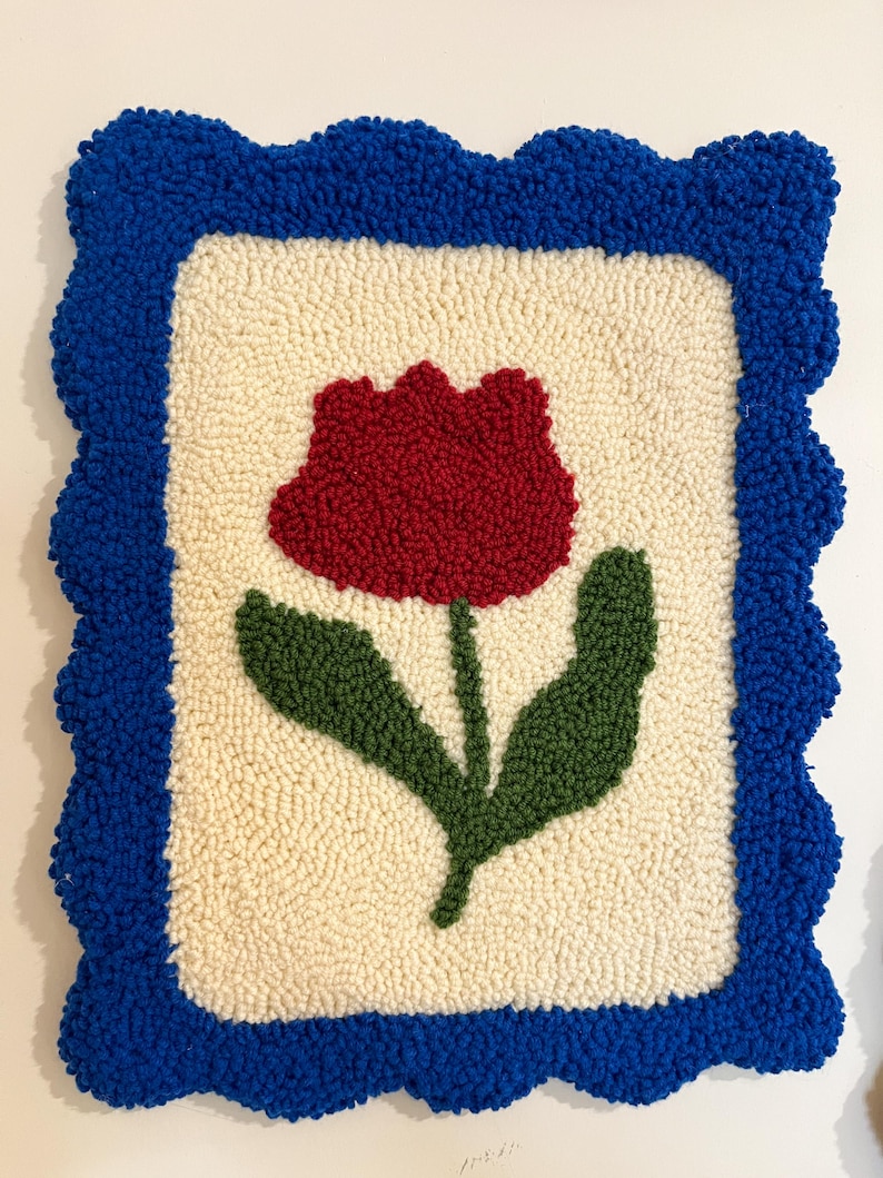 Pode incluir: Uma tape&ccedil;aria de parede texturizada feita &agrave; m&atilde;o com uma tulipa vermelha e folhas verdes sobre um fundo creme. O design &eacute; emoldurado por uma borda ondulada azul brilhante. A obra de arte &eacute; feita com uma t&eacute;cnica de fio em la&ccedil;o.