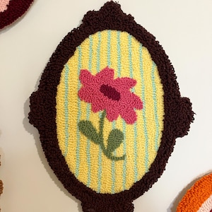 Pode incluir: Uma tape&ccedil;aria de parede oval feita &agrave; m&atilde;o com uma flor rosa e folhas verdes sobre um fundo amarelo com listras azuis. A flor &eacute; emoldurada por uma borda marrom escura texturizada. Outras pe&ccedil;as semelhantes s&atilde;o vis&iacute;veis.